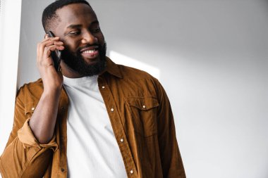 Çekici gülümseyen Afrikalı genç adam, pencerenin önünde beklerken cep telefonu kullanıyor.