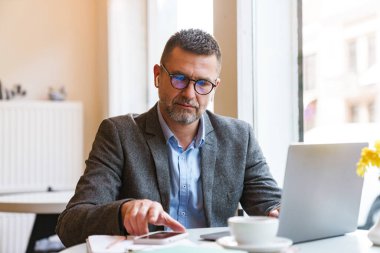 Kapalı bir kafede oturup, kahve içerken, kulaklık takarken dizüstü bilgisayarda çalışan yakışıklı, kendine güvenen bir iş adamı.