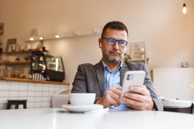 Genç adam, kulaklıkla müzik dinlerken akıllı telefondan mesaj atıyor ve kahve dükkanında ücretsiz internet bağlantısının tadını çıkarıyor.