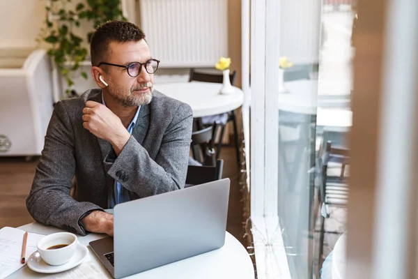 カフェに座っている間 ラップトップコンピュータで働いているハンサムな自信のあるビジネスマン コーヒーを飲む イヤホンを身に着けている — ストック写真