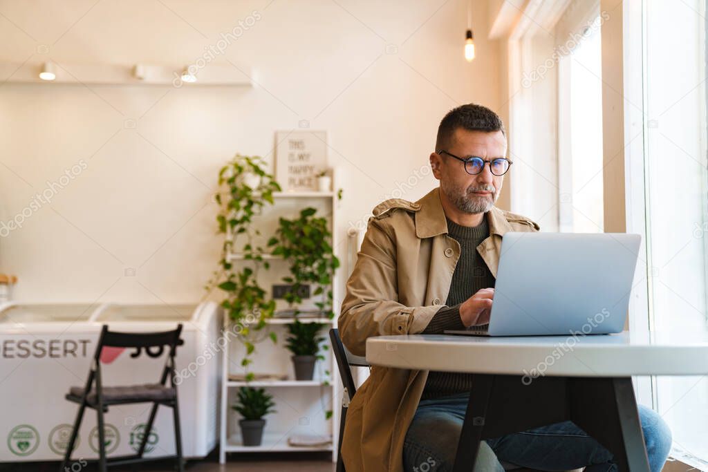 Handsome confident businessman working on laptop computer while sitting in cafe indoors, drinking coffee, wearing earphones