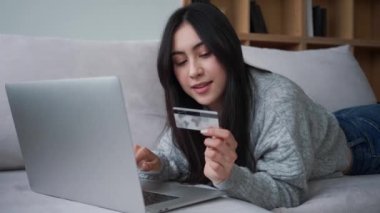 Gülümseyen genç Asyalı bir kadın elinde kredi kartıyla apartmandaki kanepede yatarken bilgisayarını kullanıyor.