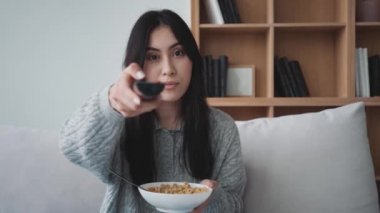 Genç ve güzel bir Asyalı kadın apartmandaki kanepede oturmuş televizyon izlerken tahıl gevreği yiyor.