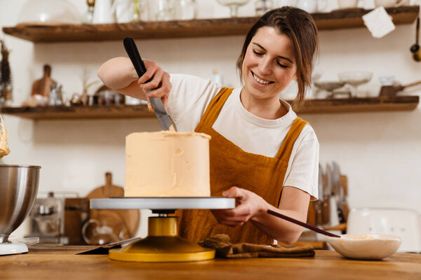 Beautiful happy pastry chef woman smiling while making cake with cream at cozy kitchen