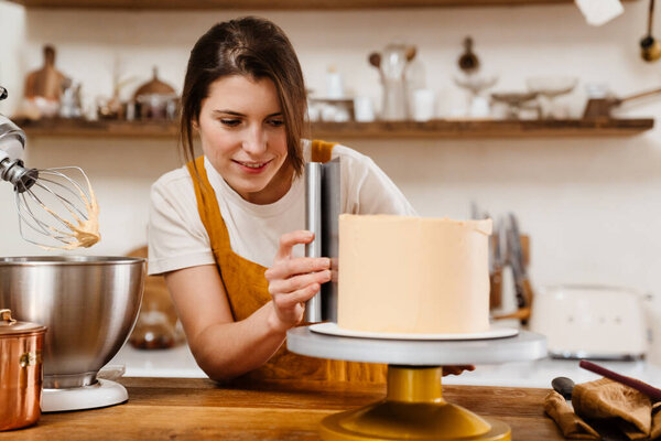 Beautiful pleased pastry chef woman making cake with cream at cozy kitchen