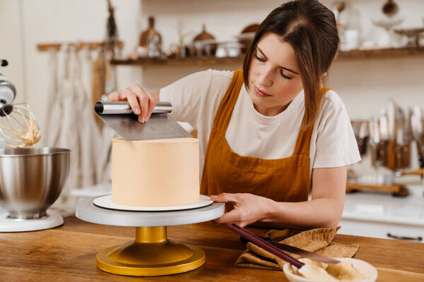 Beautiful focused pastry chef woman making cake with cream at cozy kitchen