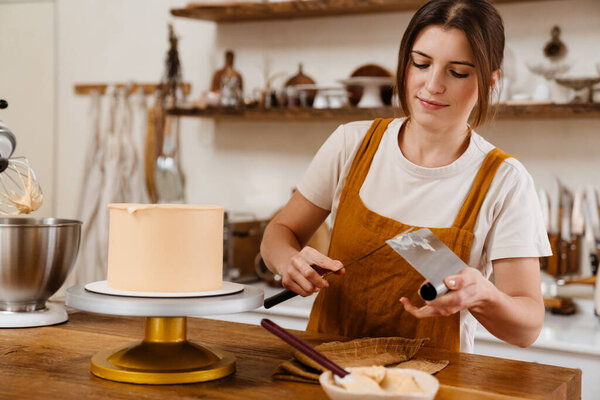Beautiful pleased pastry chef woman making cake with cream at cozy kitchen