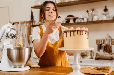 Güzel, memnun pasta şefi kadın rahat bir mutfakta cep telefonuyla pasta fotoğrafı çekiyor.