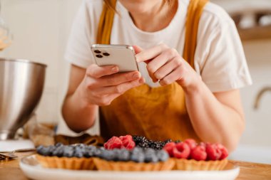 Kafkasyalı pasta şefi kadın rahat bir mutfakta cep telefonuyla tart fotoğrafı çekiyor.