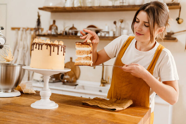 Beautiful happy pastry chef woman showing piece of cake with cream at cozy kitchen