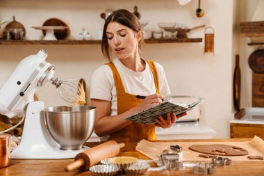 Konsantre olmuş pasta şefi kadın, sıcak mutfakta Noel kurabiyesi yaparken not yazıyor.