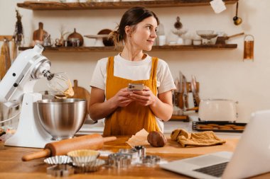 Gülümseyen pasta şefi kadın rahat mutfakta kek pişirirken cep telefonu kullanıyor.