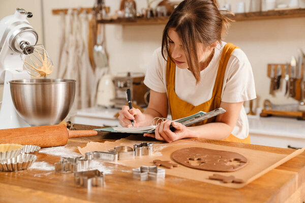 Focused pastry chef woman writing down notes while making Christmas cookies at cozy kitchen