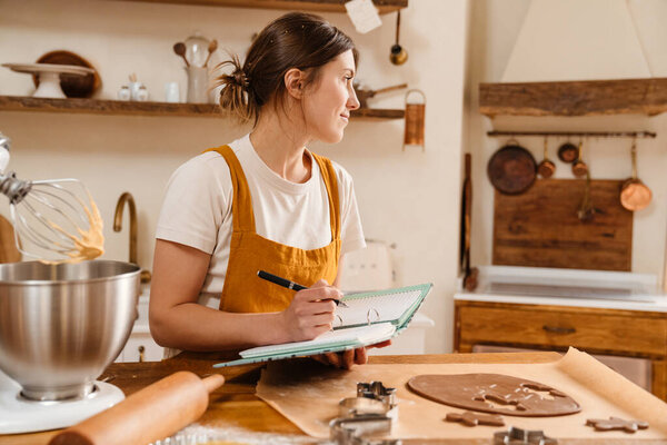 Pleased pastry chef woman writing down notes while making Christmas cookies at cozy kitchen