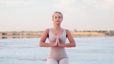 A calm young blonde woman is doing yoga holding hands in a praying gesture sitting on the yoga mat outside on nature