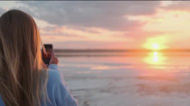 A back-view of a young blonde woman is taking photo of a sun using her mobile phone while walking outside on nature