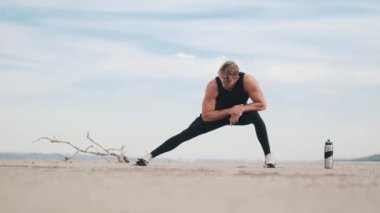 A muscular young sportsman is doing stretching exercises on his legs while training outdoors