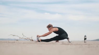 A good-looking young sportsman is doing stretching exercises on his legs while training outdoors