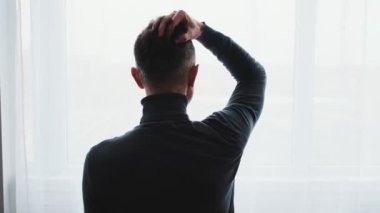 A back view of a mature man in a gray turtleneck looking out the window and fixing his hair