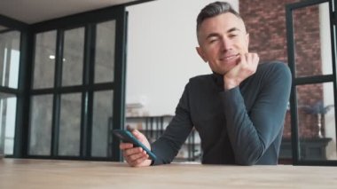 A thoughtful man siting at the table and looking out the window while holding the phone in his hands on the background of the office