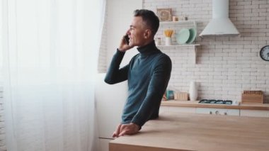 A man standing and talking on the phone in a green turtleneck in the background of the kitchen