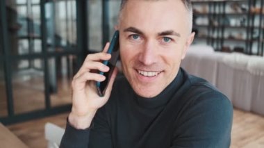 The smiling man in a green turtleneck talking on the phone, but then starting to deny something in a bright room