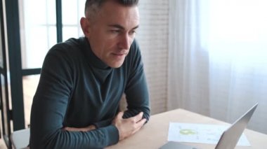 A side view of a mature man sitting at a desk in front of a monitor, talking emotionally over a video connection