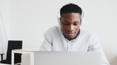 A happy black guy with headphones is doing winner gesture using his laptop while sitting in the office