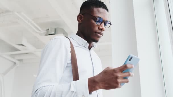 Happy Black Guy Wearing Eyeglasses Using His Smartphone Standing Window — Stockvideo