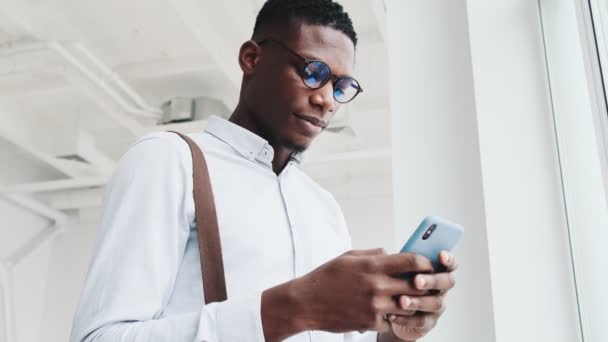 Smiling Black Guy Wearing Eyeglasses Using His Smartphone Standing Window — Vídeo de Stock