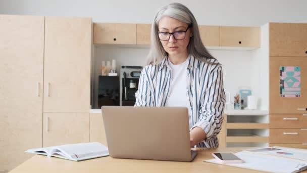 自宅のキッチンで働いている間 眼鏡をかけた重大な高齢のビジネス女性がノートパソコンを使用しています — ストック動画