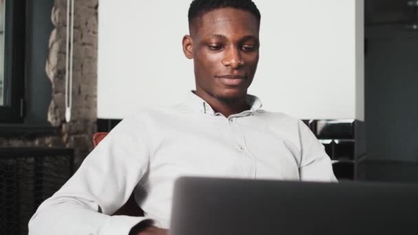 一位面带微笑的非洲裔美国人正在使用笔记本电脑 他坐在阁楼公寓里的椅子上 — 图库视频影像