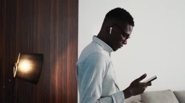 A side-view of a handsome african american businessman wearing wireless earphones is using his smartphone standing in the loft apartments