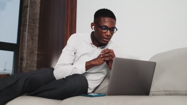 一位戴着无线耳机的快乐的非洲商人正在和他的笔记本电脑一起工作 他坐在阁楼公寓的沙发上 在智能手机上得到了一个很好的信息 — 图库视频影像