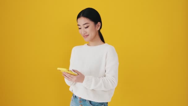 Drömmande Asiatisk Kvinna Använder Sin Smartphone Stående Isolerad Över Gul — Stockvideo
