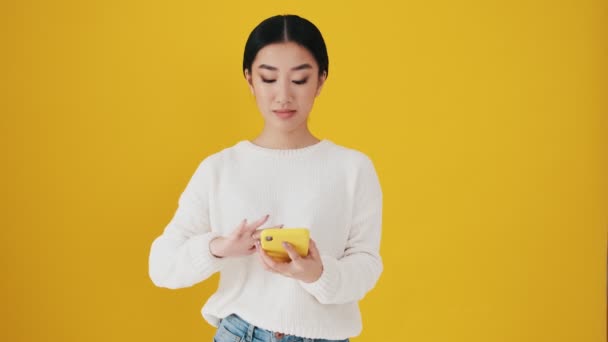 Besviken Asiatisk Kvinna Sveper Sin Smartphone Stående Isolerad Över Gul — Stockvideo