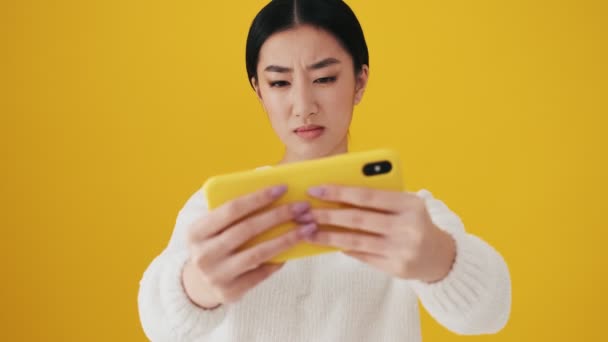 スタジオで黄色の壁の上に隔離された彼女のスマートフォンにゲームをプレイしている深刻なアジアの女性 — ストック動画