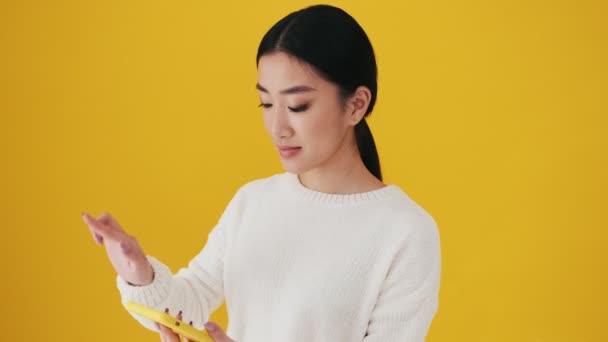 スタジオで黄色の壁の上に隔離された立って勝者ジェスチャーをしながら幸せなアジアの女性は彼女のスマートフォンで話しています — ストック動画