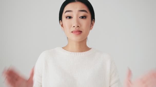白いセーターを身に着けているかなり素敵なアジアの女性は何かを求めています彼女の手で祈るジェスチャーで立っている間に白い壁の上に隔離されたスタジオ — ストック動画