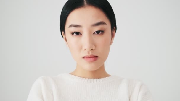 白いセーターを着たアジア系の女性のクローズアップは スタジオの白い壁の上に隔離されたカメラスタンドを探しています — ストック動画
