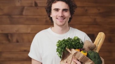 The smiling man standing with groceries in paper bag and showing okay while standing in the wooden studio