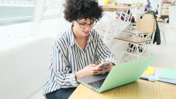 カフェに座っているノートパソコンを操作している女性がスマホを使っている — ストック動画