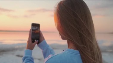 A back-view of a young blonde woman is taking photo of a dawn using her smartphone walking outside on nature