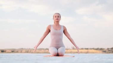A beautiful young blonde woman with closed-eyes is doing yoga holding hands in a praying gesture sitting on the yoga mat outside on nature