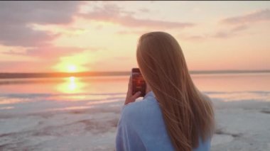 A back-view of a young blonde woman is taking photo of a sun using her smartphone walking outside on nature