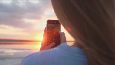 A close-up view of a young blonde woman is taking photo of a sun using her mobile phone while walking outside on nature