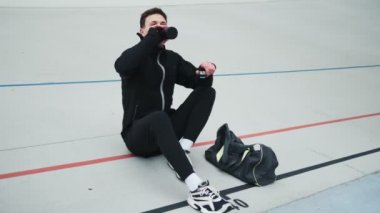 Yakışıklı genç bir sporcu spor sahasında otururken bir spor şişesinden su içiyor.
