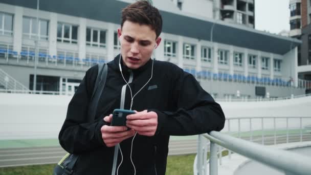 トレーニングの後 スタジアムに立っている間 焦点を当てた若いランナーがスマートフォンを使用しています — ストック動画