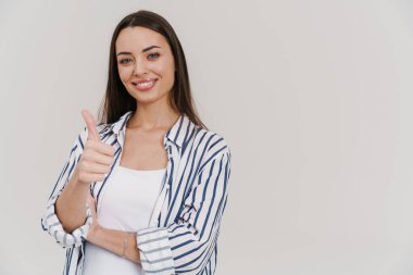 Mutlu, güzel, esmer kız gülümseyen ve baş parmağını beyaz arka planda gösteren.
