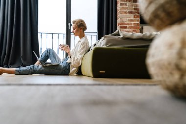 Evdeki kanepede yatarken dizüstü bilgisayardan gülümseyen genç bir kadın.